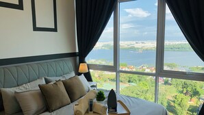 A hotel room with a large bed, a bedside table with a lamp, and a view of a lake and cityscape. - Paragon Suite By Antlerzone (Johor Bahru)