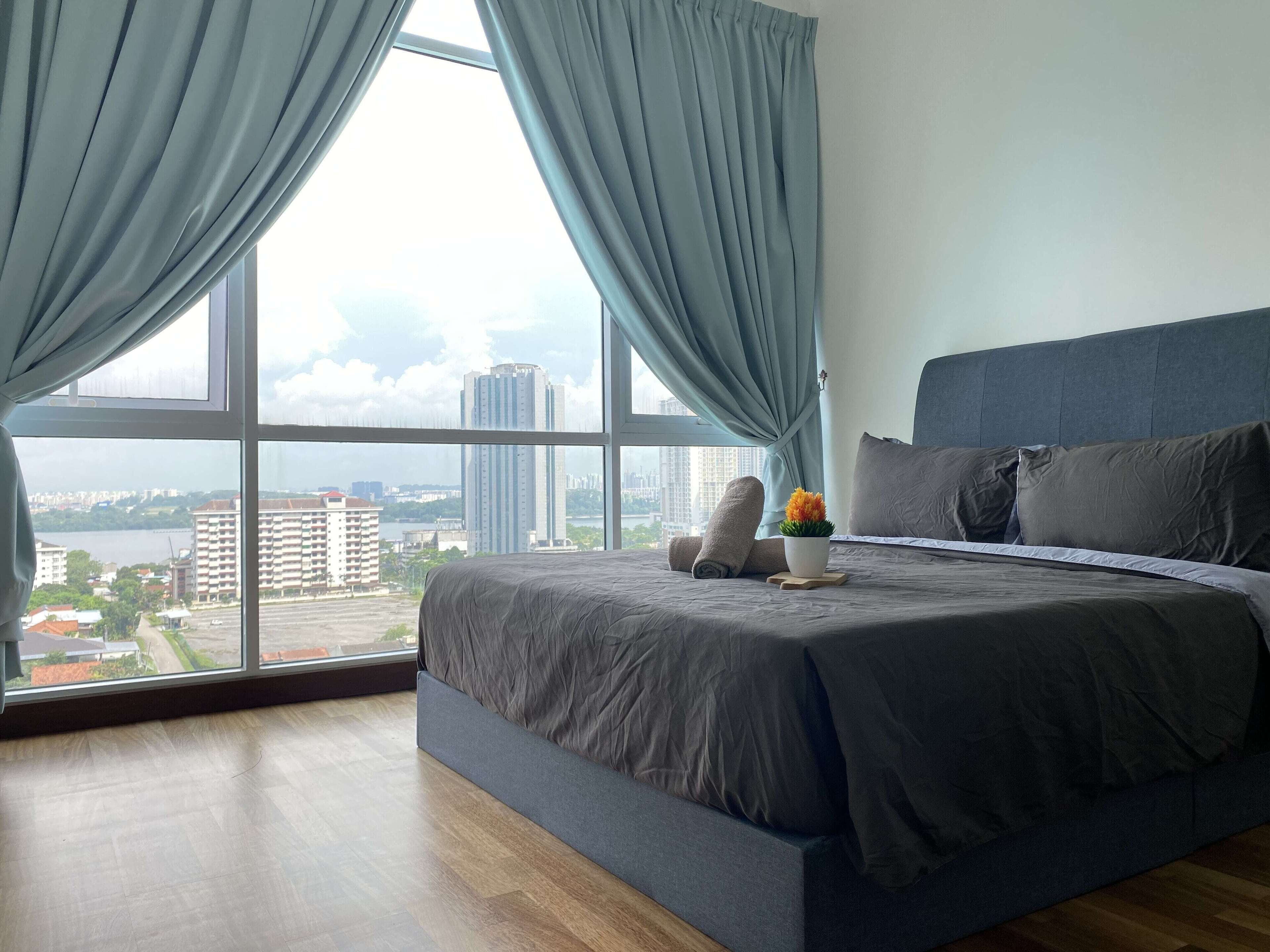 A modern bedroom with a large bed, grey bedding, and a view of a cityscape through floor-to-ceiling windows.