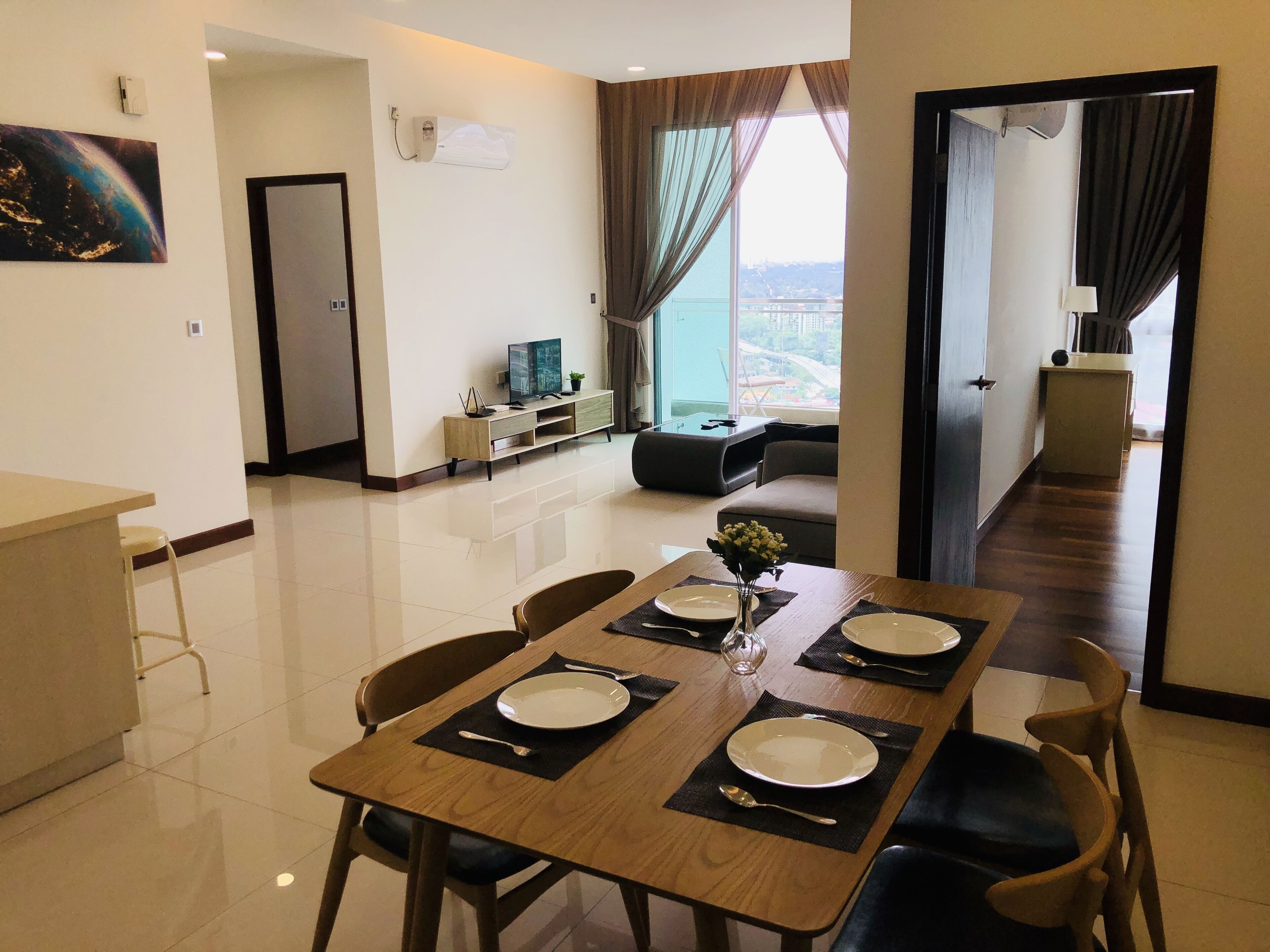 Un comedor con una mesa lista para cuatro personas, un televisor al fondo y vistas de la ciudad a través de amplios ventanales.