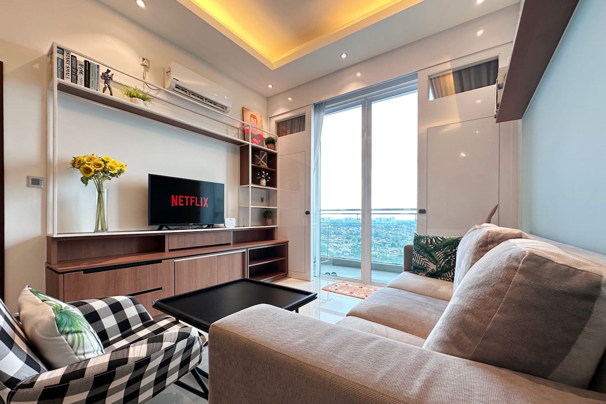 A modern living room with a flat-screen TV, a sofa, a coffee table, and a balcony with a city view.