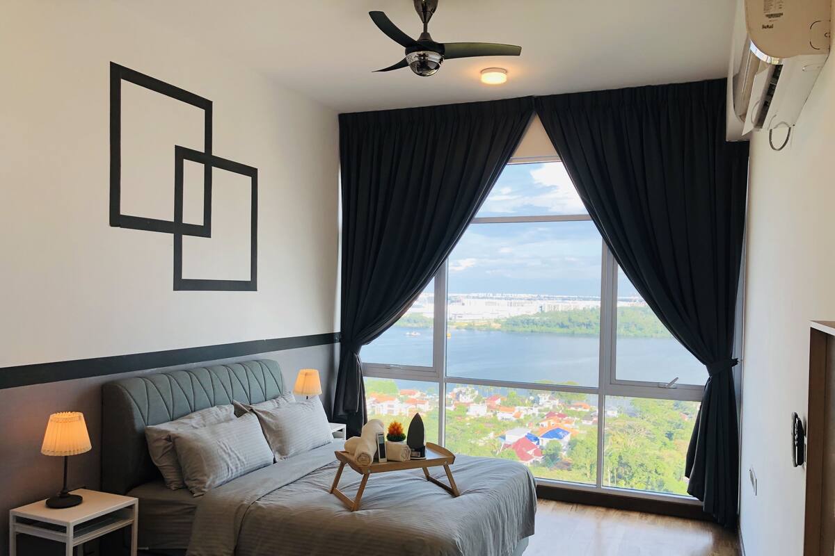 A bedroom with a bed, a nightstand, a lamp, a window with curtains, and a view of a body of water and buildings.