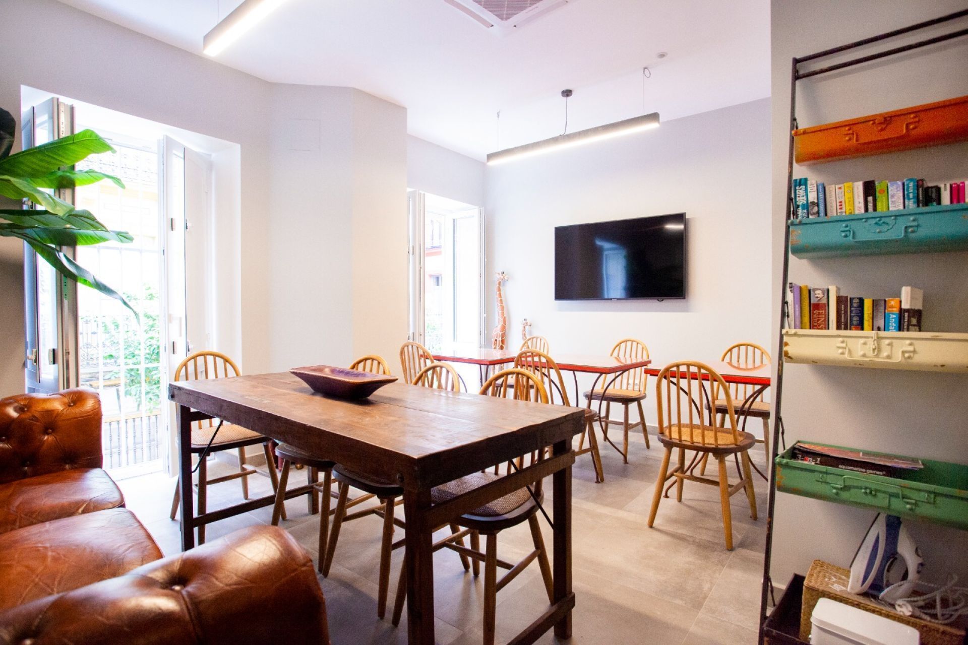 Un comedor con una mesa y sillas de madera, un televisor montado en la pared y una estantería con libros.