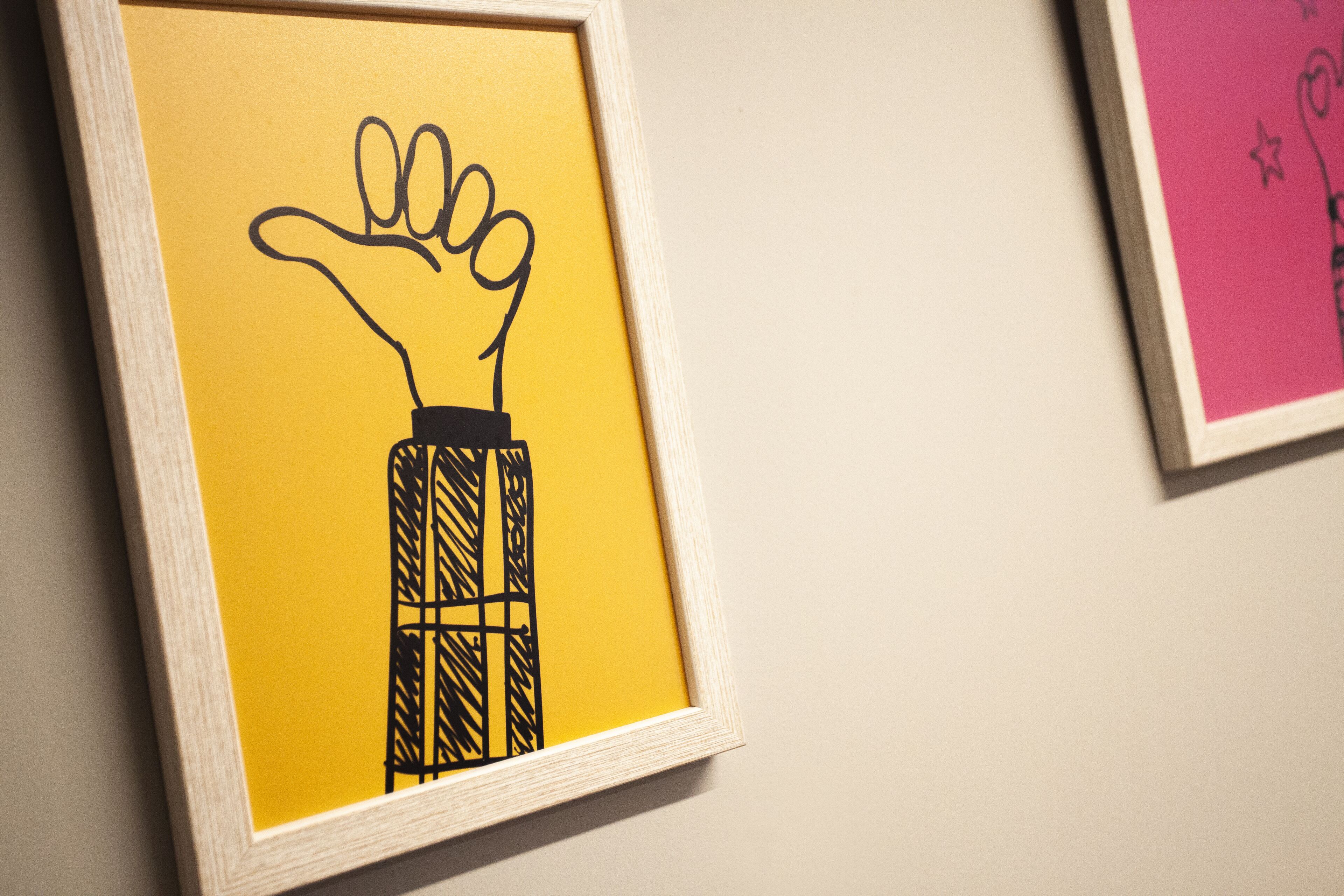 Obra de arte en marco que muestra una mano extendiéndose hacia arriba, sobre un fondo amarillo.