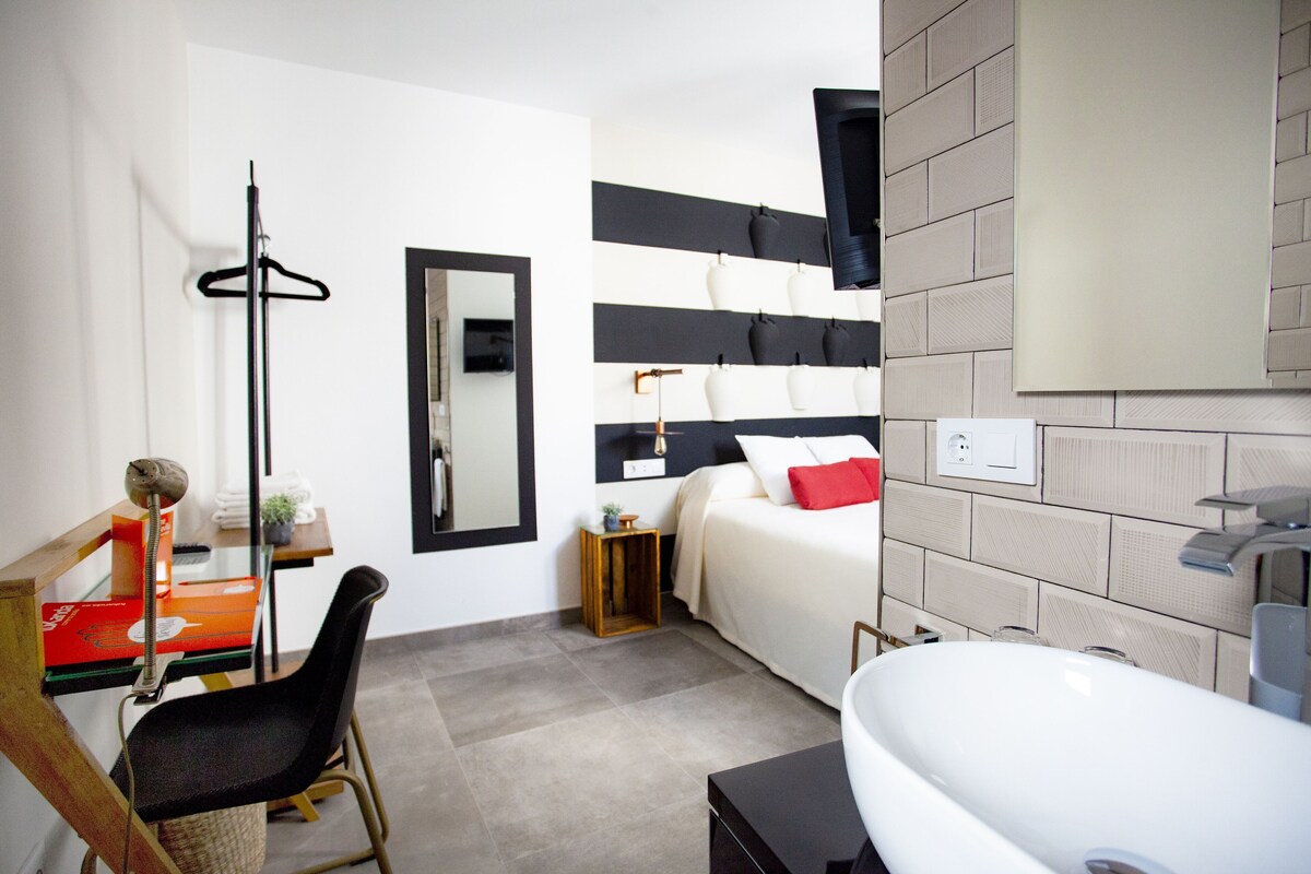 A modern hotel room with a bed, a desk, a chair, a large mirror, and a bathroom with a sink and mirror.