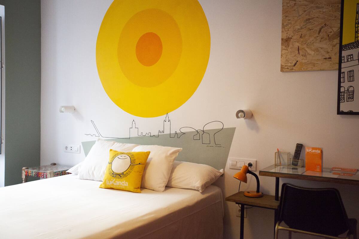 Hotellihuoneessa on sänky, työpöytä ja tuoli. Seinällä on keltainen koriste ja kaupunkimaisemaa esittävä seinämaalaus.