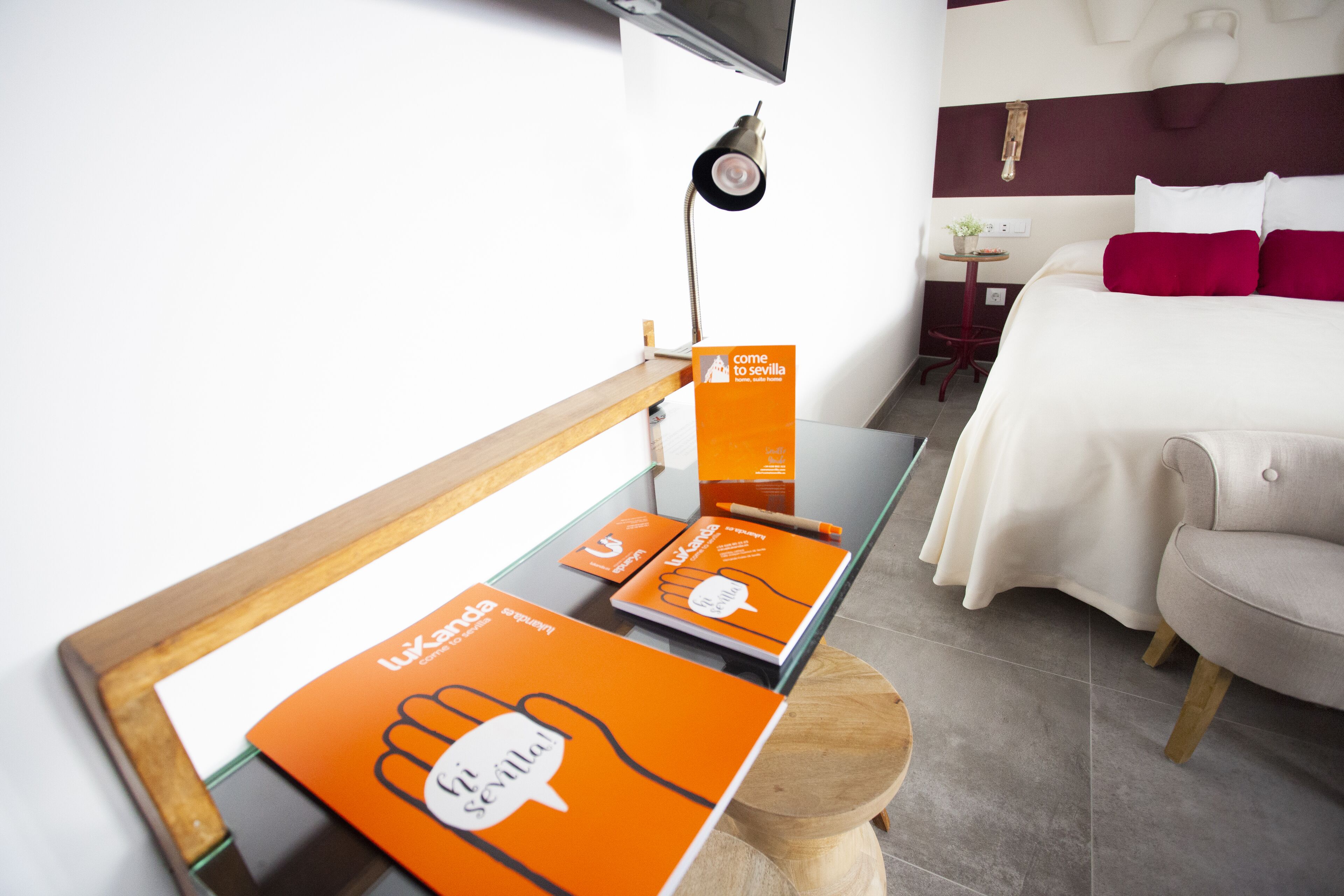 Habitación de hotel con cama, escritorio con material promocional, televisión y una pequeña mesa con una planta.