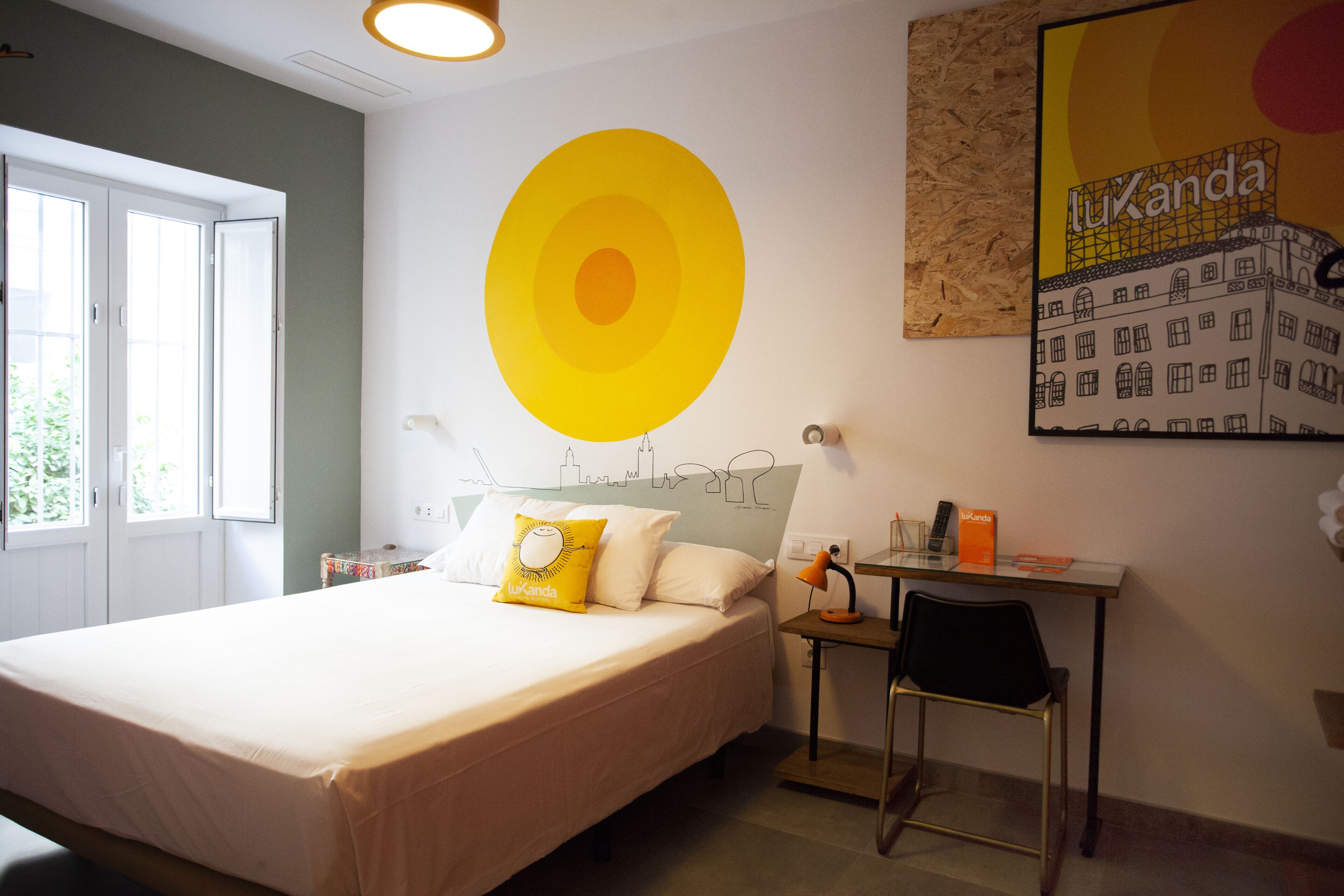 A hotel room with a bed, a desk, and a chair. There is a window with a view of greenery outside.