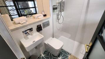 A modern bathroom with a white sink, toilet, and shower. There is a mirror above the sink and a shelf with toiletries. The floor has a geometric tile pattern.
