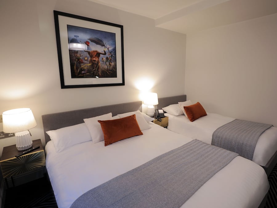 Una camera d'albergo con due letti, ognuno con lenzuola bianche e una coperta grigia. Sono presenti due lampade da comodino, un quadro appeso al muro e un comodino con un telecomando.