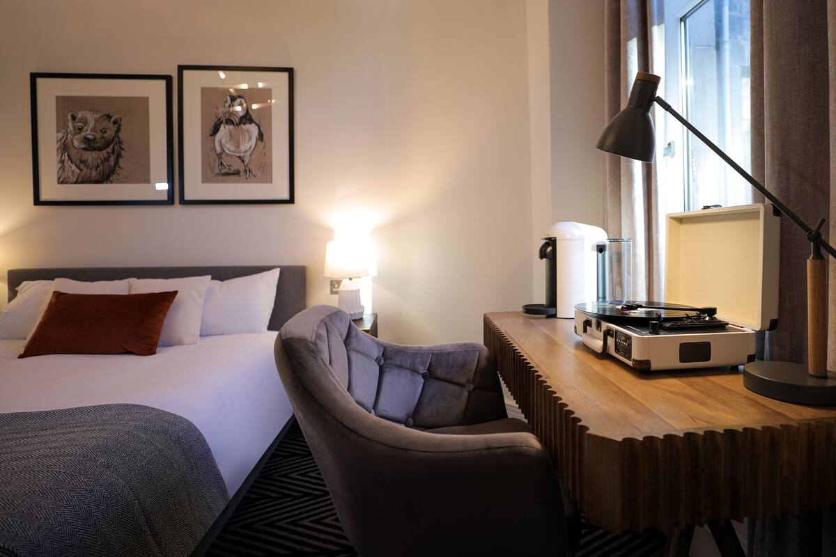 A hotel room with a bed, a desk with a turntable, a chair, and two framed pictures on the wall.