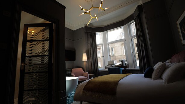 A hotel room with a large bed, a desk with a lamp, a chair, and a view of the city through large windows. - Boutique 50 (Glasgow)