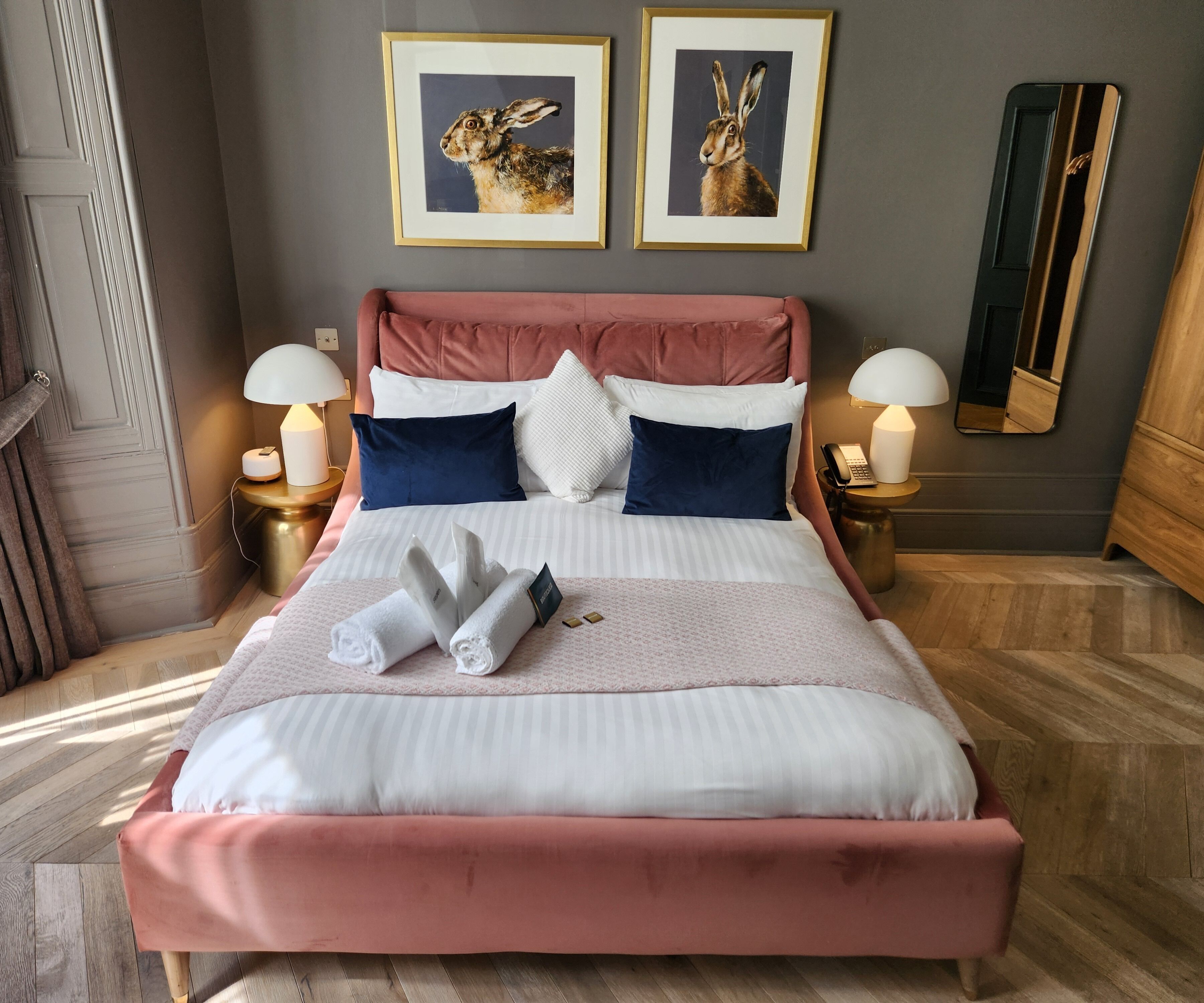 Una cama bien hecha con ropa de cama rosa y blanca, dos cuadros en la pared y una mesita de noche con una lámpara y teléfono.