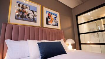 A hotel room with a large bed, a headboard, two framed artworks on the wall, and a large window.