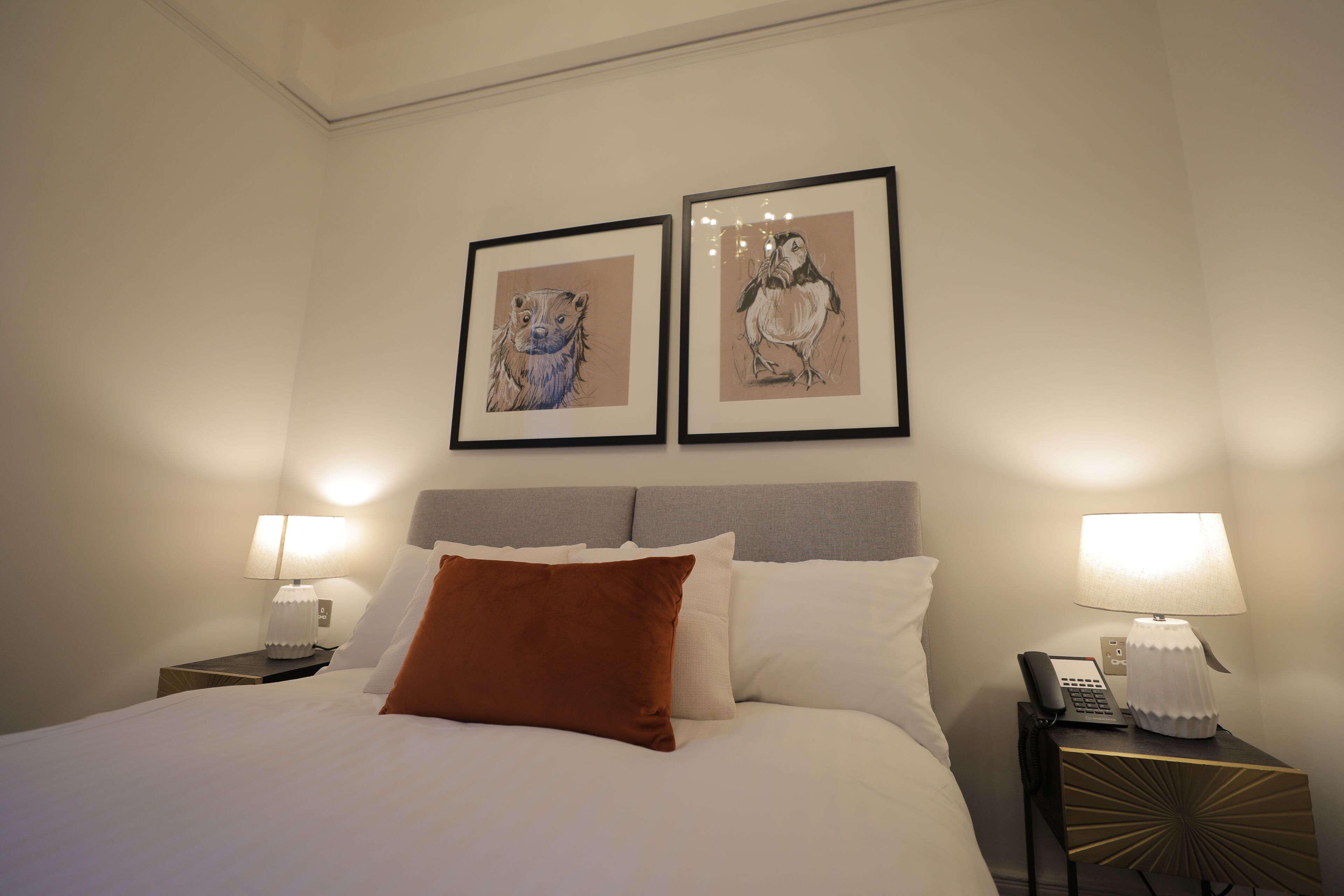 Una habitación de hotel con cama, dos ilustraciones enmarcadas de animales en la pared, una mesita de noche con lámpara y teléfono.
