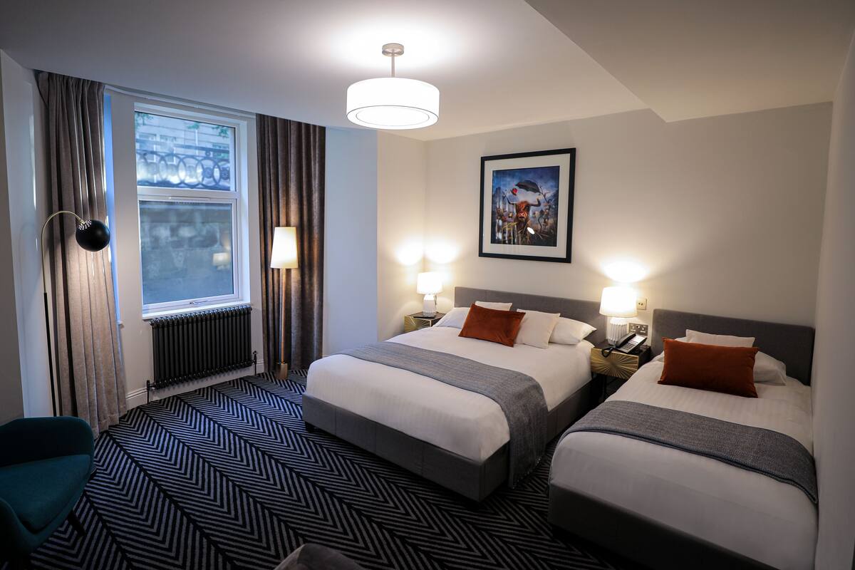 A hotel room with two beds, a large window with curtains, a radiator, a desk with a lamp, and a painting on the wall.
