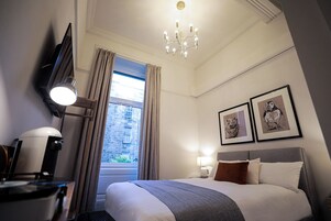 A hotel room with a bed, a desk, a chair, a lamp, a chandelier, and two framed pictures on the wall.