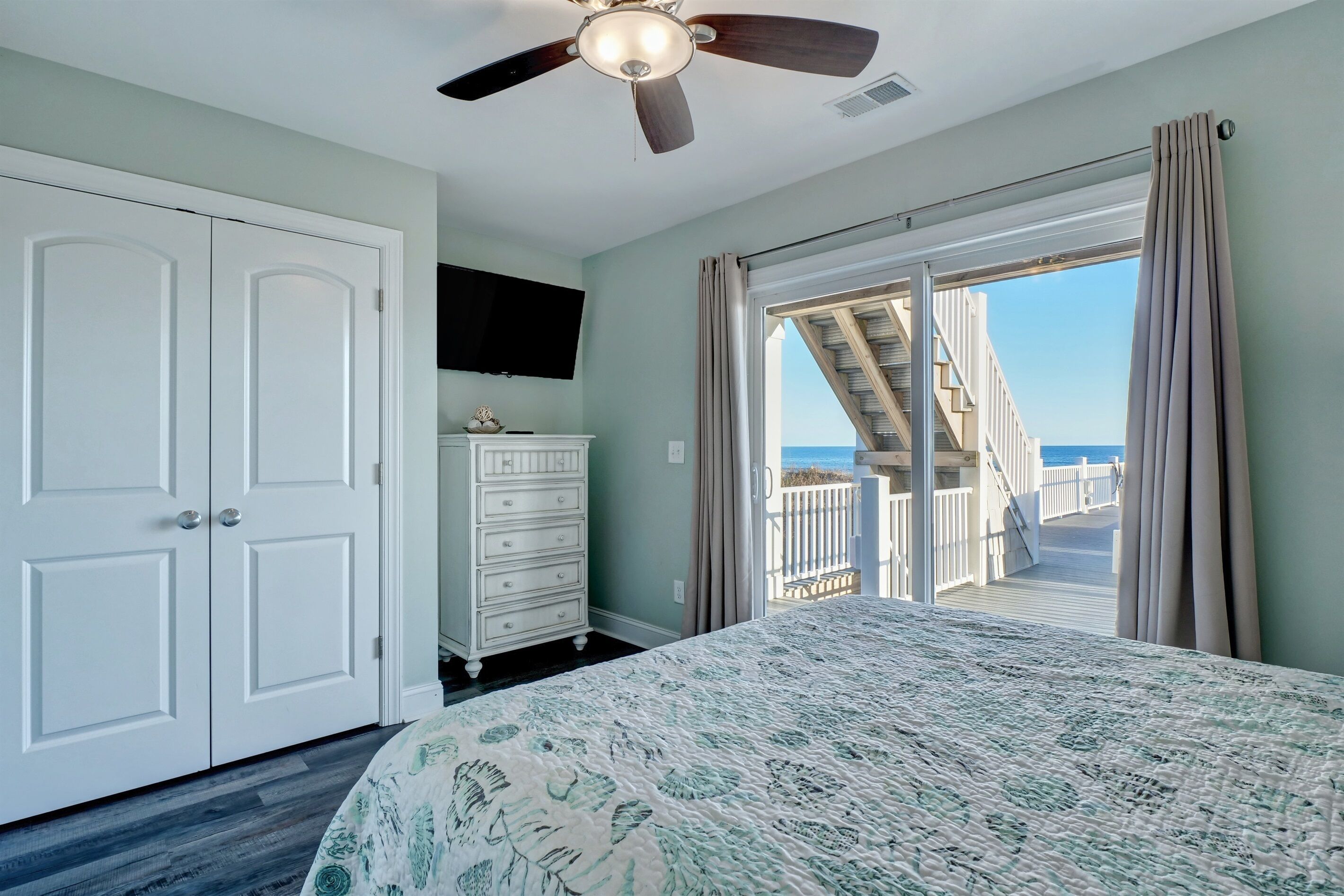 A bedroom with a bed, a dresser, a ceiling fan, and a view of the ocean through a sliding glass door.