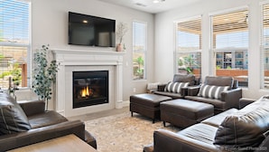 A modern living room with a fireplace, leather sofas, a flat-screen TV, and large windows with blinds.