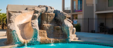 A swimming pool with a rock waterfall feature, surrounded by a modern building with balconies.