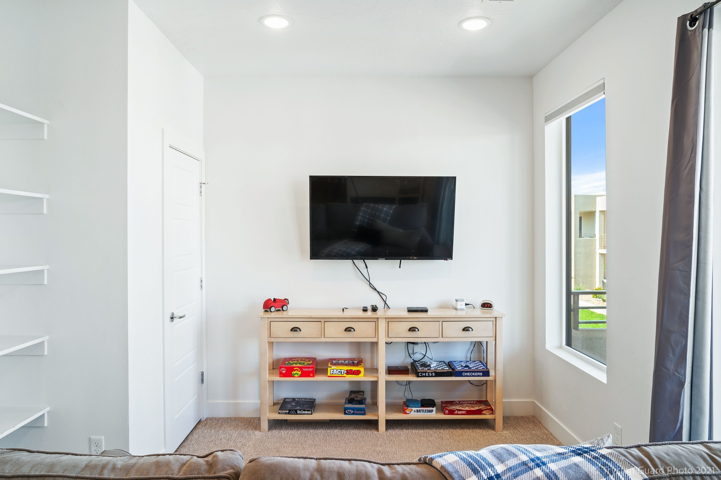 A bedroom with a bed, a TV mounted on the wall, a wooden dresser with drawers, and a window with curtains.