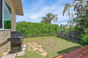 Un jardin avec un barbecue, un chemin en pierre et une végétation luxuriante comprenant une haie et des palmiers.