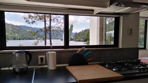 Uma cozinha com uma grande janela dando para um lago e montanhas, fogão a gás, tábua de corte e chaleira.