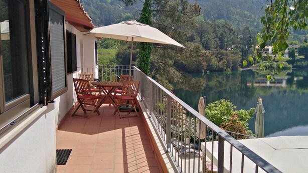 Uma varanda com mesa e cadeiras, um grande guarda-sol e vista para um lago rodeado por vegetação.