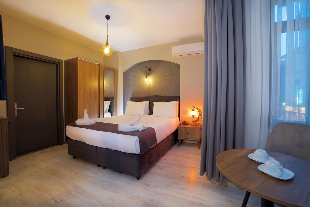 A hotel room with a large bed, a bedside table with a lamp, a chair, and a view of the outside through a window with curtains.