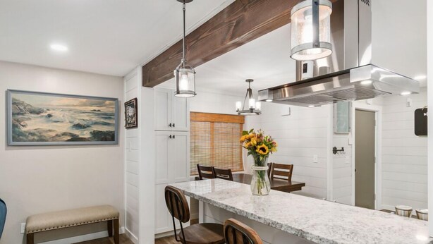 Una cocina con encimera de mármol, sillas de madera, un banco, lámparas colgantes y un cuadro grande en la pared.