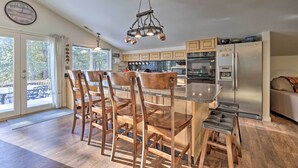 A modern kitchen with wooden cabinets, a stainless steel refrigerator, and a dining area with wooden chairs and a bar stool. - 'Peaceful Pines' - Cascade Cabin w/ Hot Tub! (Cascade)