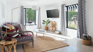 Una sala de estar moderna con un sofá, una mesa de centro y un televisor de pantalla plana. Hay ventanas amplias con cortinas estampadas, un ventilador de techo y una estantería.