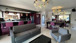 A modern living room with a grey sofa, a coffee table, and a dining area with purple chairs. There is a kitchen in the background with a black refrigerator and a chandelier above the dining table. - Villa Naeva with swimming pool, Jacuzzi 10 places, Ping pong, plancha and Brasero (Agnac)