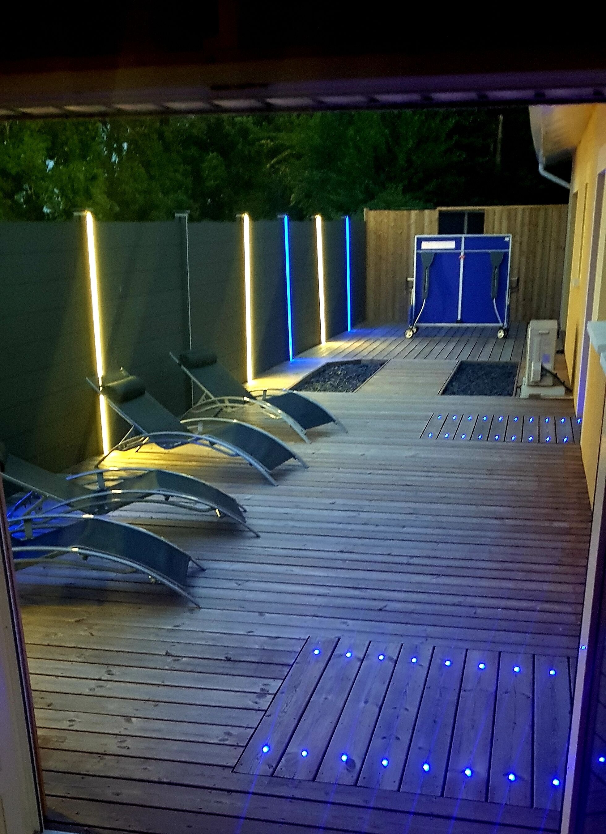 Een houten terras met ligbedden en een blauw verlichte muur.