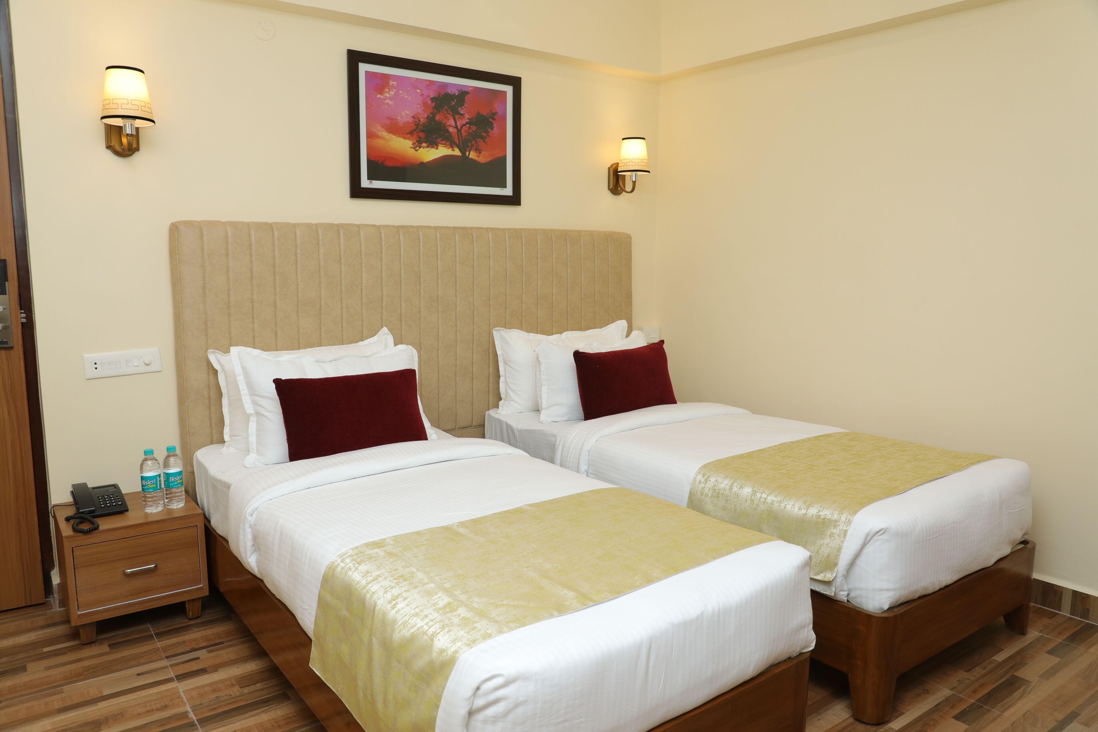 A hotel room with two beds, a nightstand with a phone and water bottles, and a painting on the wall.