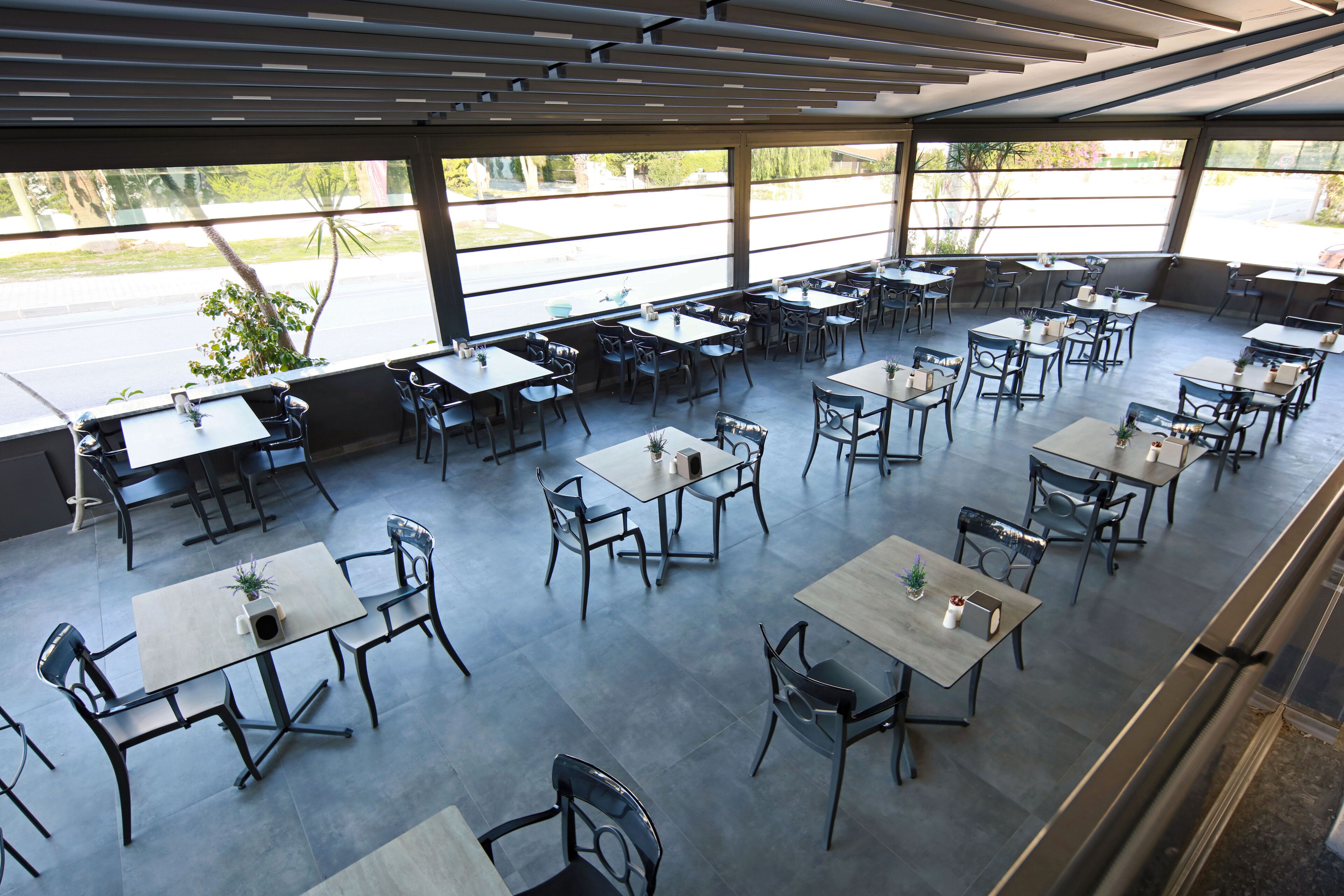 A spacious outdoor dining area with multiple tables and chairs, large windows offering views of the street and greenery.