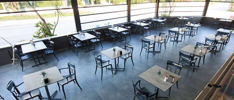 A spacious outdoor dining area with multiple tables and chairs, large windows offering views of the street and greenery.