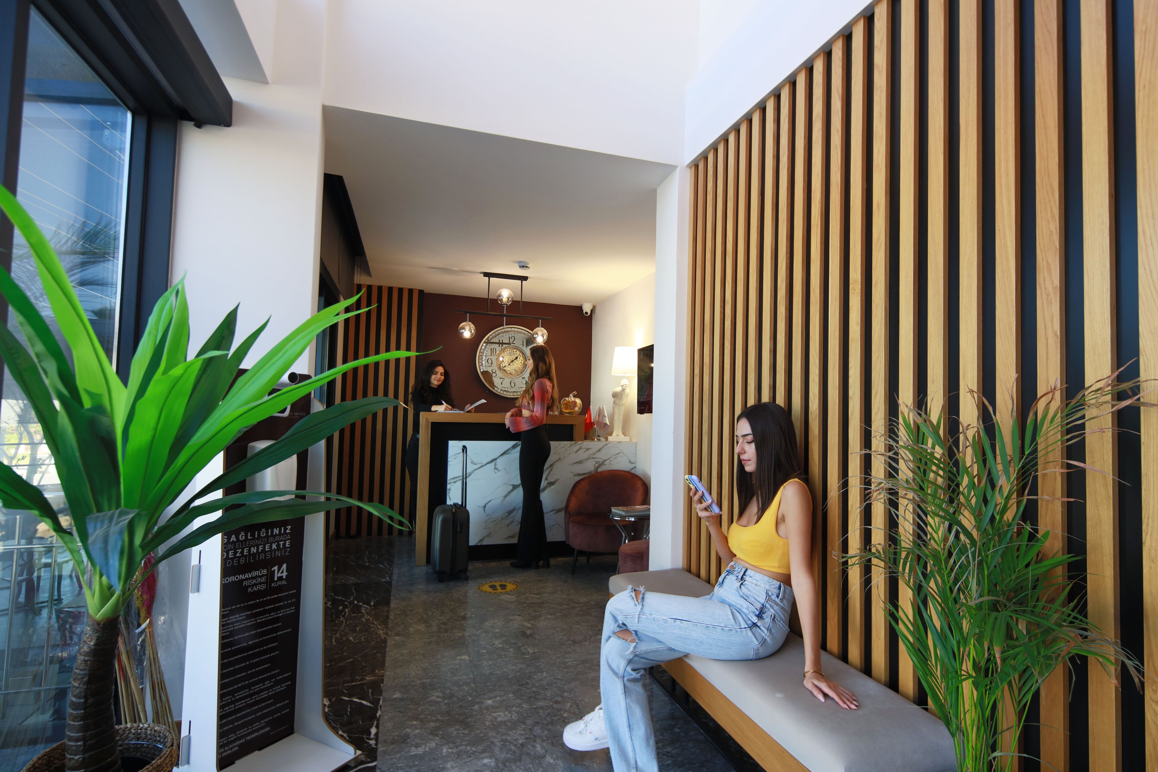Una mujer sentada en un banco en un vestíbulo moderno con una mesa de recepción, un reloj y una planta.