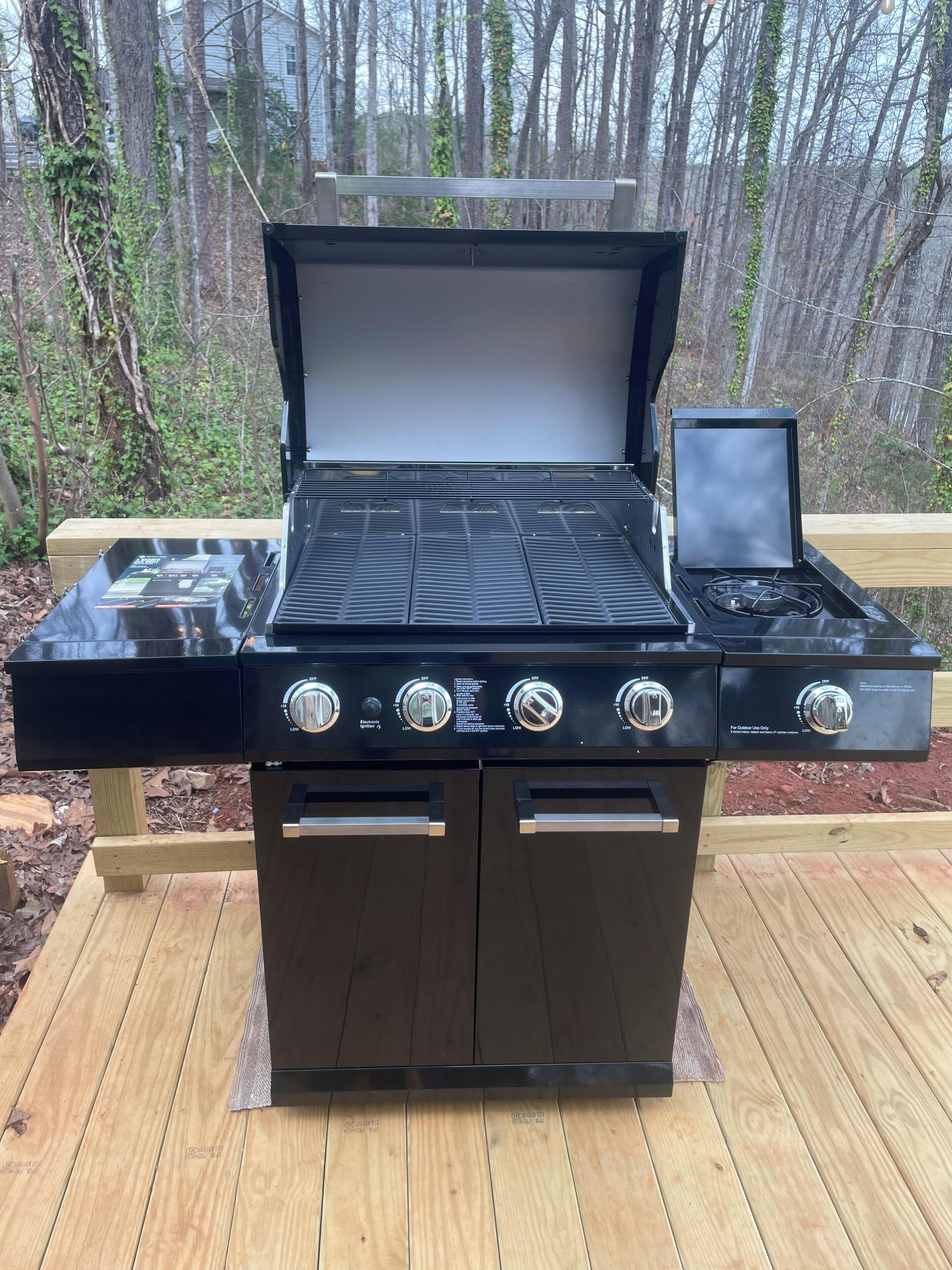 A black outdoor grill with four burners and a digital display, set on a wooden deck with a forest backdrop.