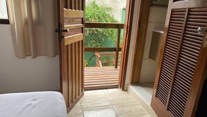 Un couloir étroit, avec un lit sur la gauche, une porte en bois à panneaux ajourés sur la droite, et une vue sur un balcon doté d’une balustrade en bois.