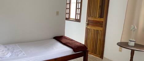 A single bed with a wooden frame and a white mattress, a wooden door with a window, and a small round table with a lamp.