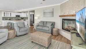 A modern living room with a fireplace, grey sofas, a wicker ottoman, and a kitchen in the background.