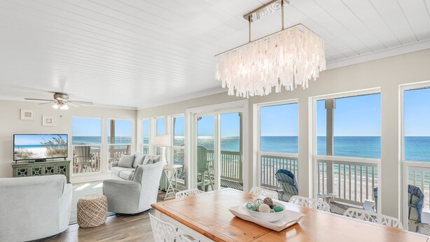 A spacious living area with a wooden dining table, white chairs, and a view of the ocean through large windows.