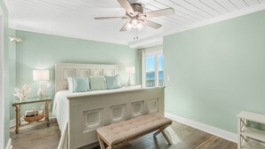 A bedroom with a bed, a bench, a ceiling fan, a nightstand, and a window with a view.