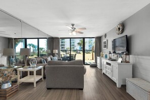 A spacious living room with a grey sofa, a white coffee table, and a TV on a white cabinet. Large windows offer a view of a balcony and palm trees.