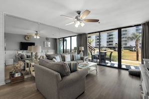 A modern living room with a grey sofa, a coffee table, and a ceiling fan. There is a large window offering a view of a building and a palm tree.
