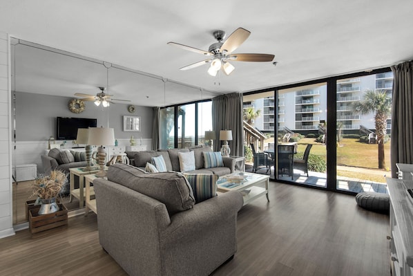 A modern living room with a grey sofa, a coffee table, and a ceiling fan. There is a large window offering a view of a building and a palm tree.