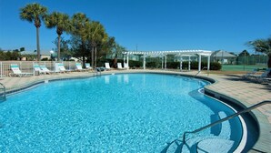 Una piscina cristalina con sillas de descanso y una zona cubierta, rodeada de palmeras y una cancha de tenis.