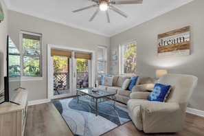 A living room with a sofa, coffee table, and a ceiling fan. There are windows with blinds and a view of the outdoors.