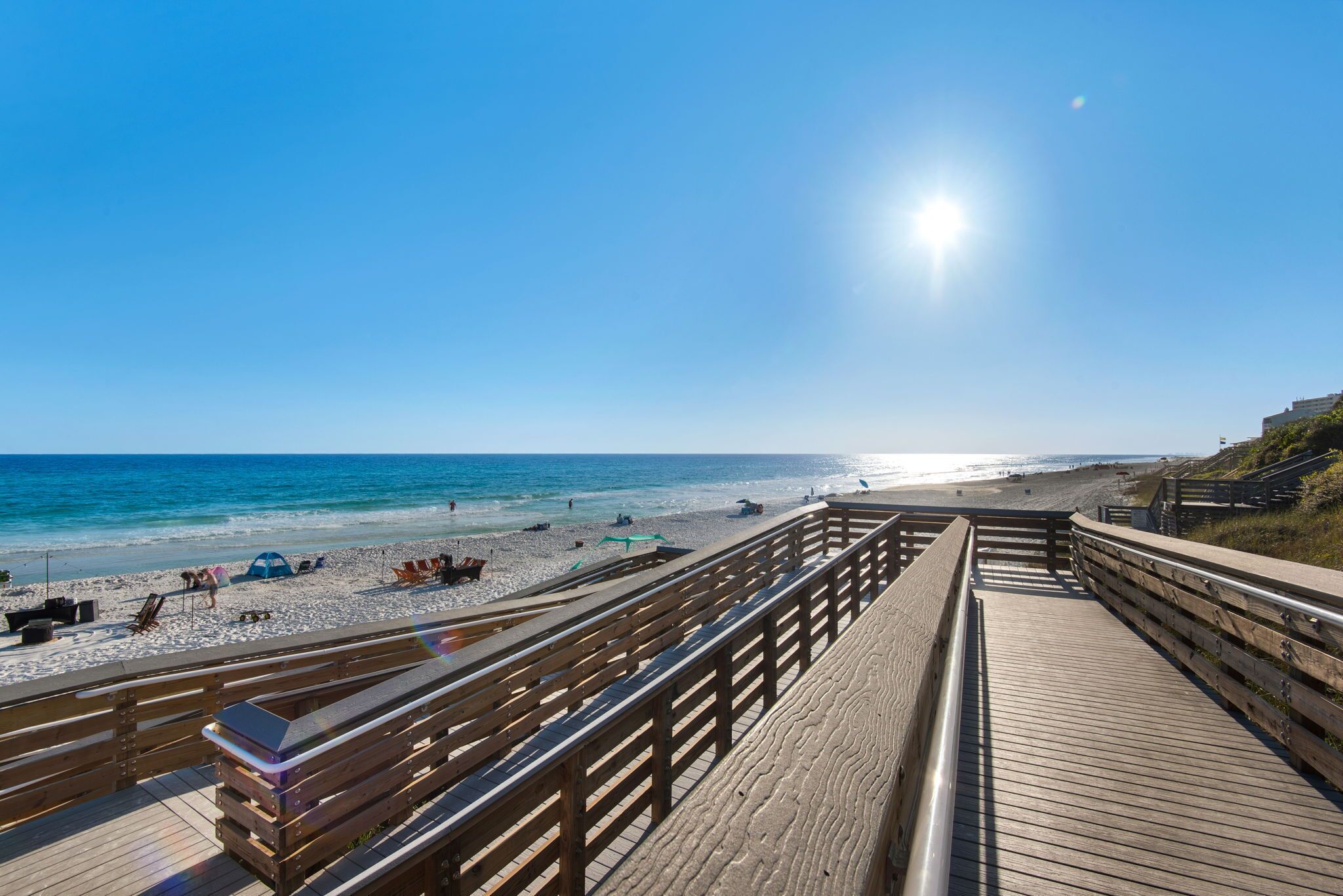 Une promenade en bois qui mène à une plage, avec un ciel dégagé et un soleil éclatant.