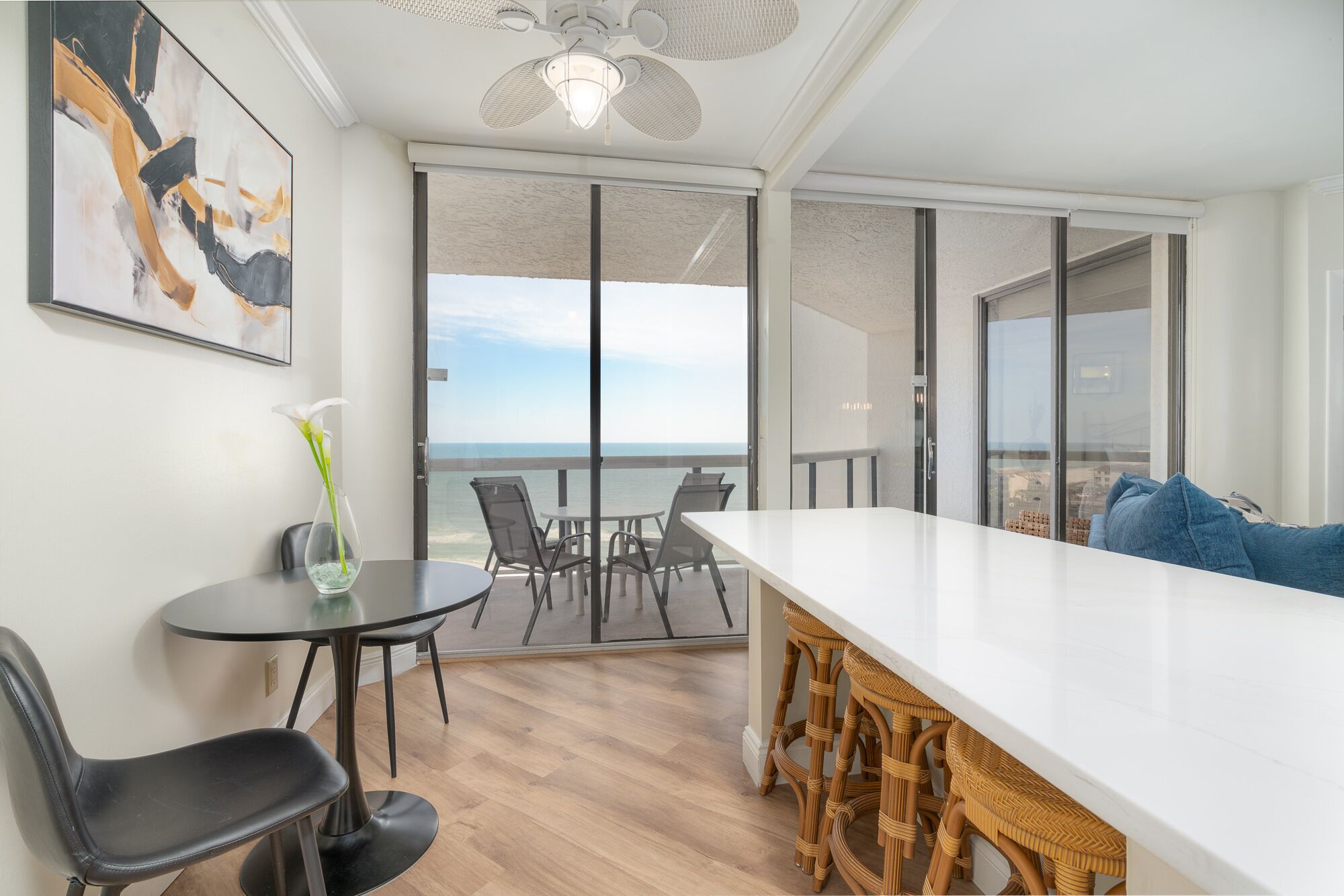 A modern room with a large white table, black chairs, and a wicker bar stool. It features a large window with a view of the ocean.